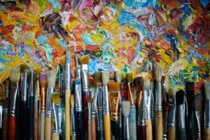op view of rows of paintbrushes laying on abstract colorful oil paint artwork.
