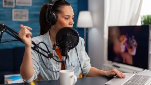 Woman wearing headphones behind microphone in a home studio. She sits at a desk with a laptop in front of her as well as white mug.
